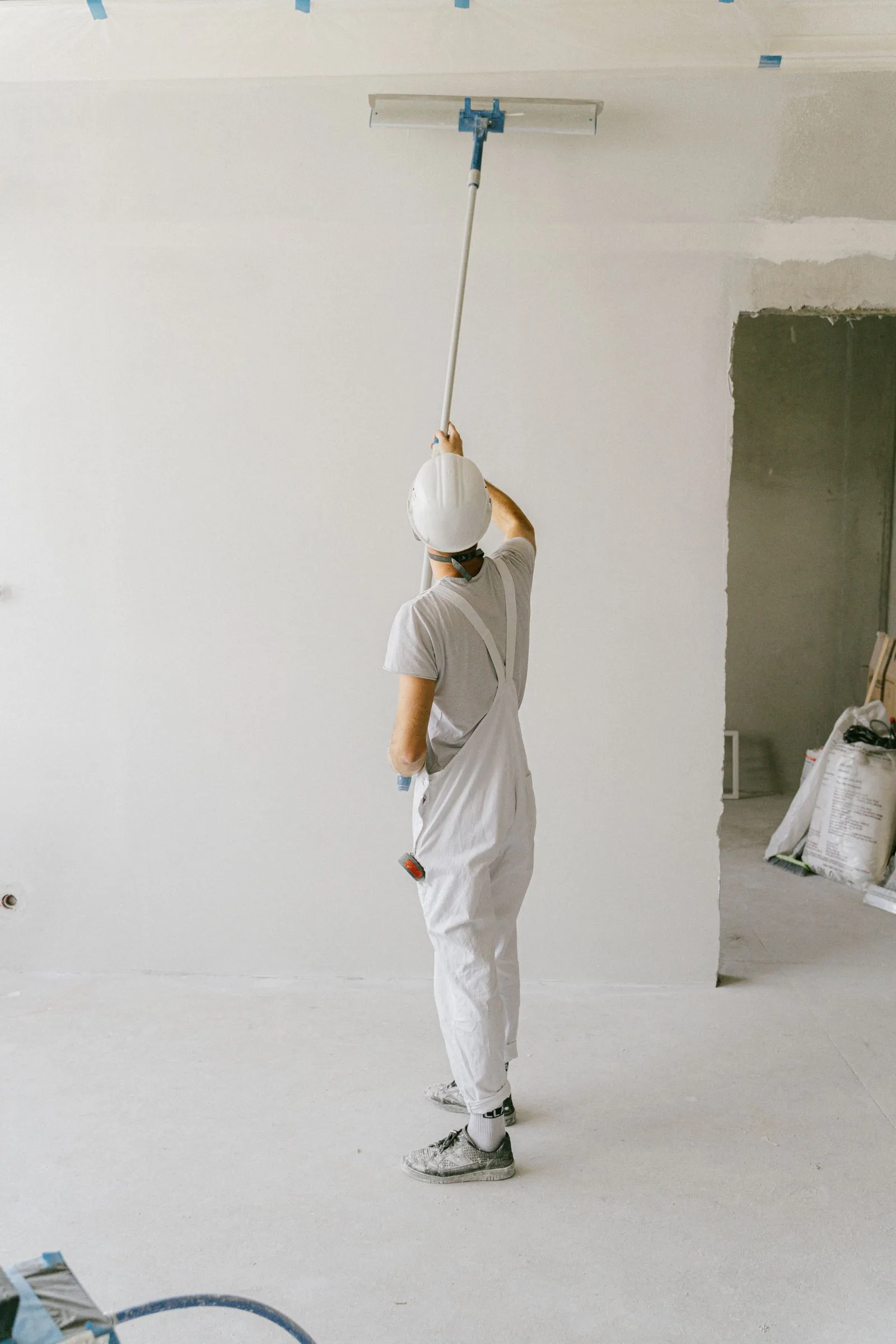 Painter in overalls and hard hat painting an interior wall with a roller