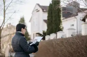 Home inspector in a winter jacket checking a floor plan on a clipboard in front of a white house