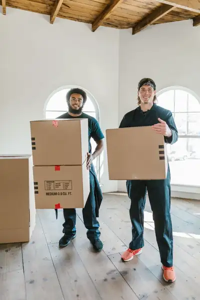 Two smiling movers in dark uniforms carrying cardboard boxes through a bright empty room