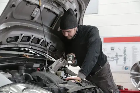 Mechanic in a beanie inspecting a car engine with a flashlight in a repair shop