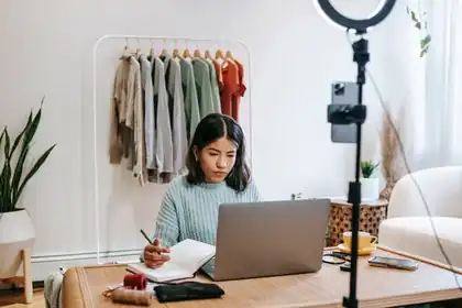 Shop owner writing notes at a laptop beside a rail of clothes and a ring light